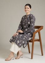 Seated model wearing Evening Meadow 2 piece printed lawn co-ord set by Mirasl with charcoal floral shirt, off-white trouser, and summer ready styling.