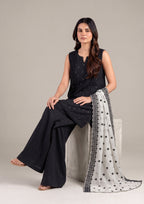 Woman sitting gracefully wearing a black embroidered sleeveless kurta with black palazzo pants and a white embroidered dupatta.