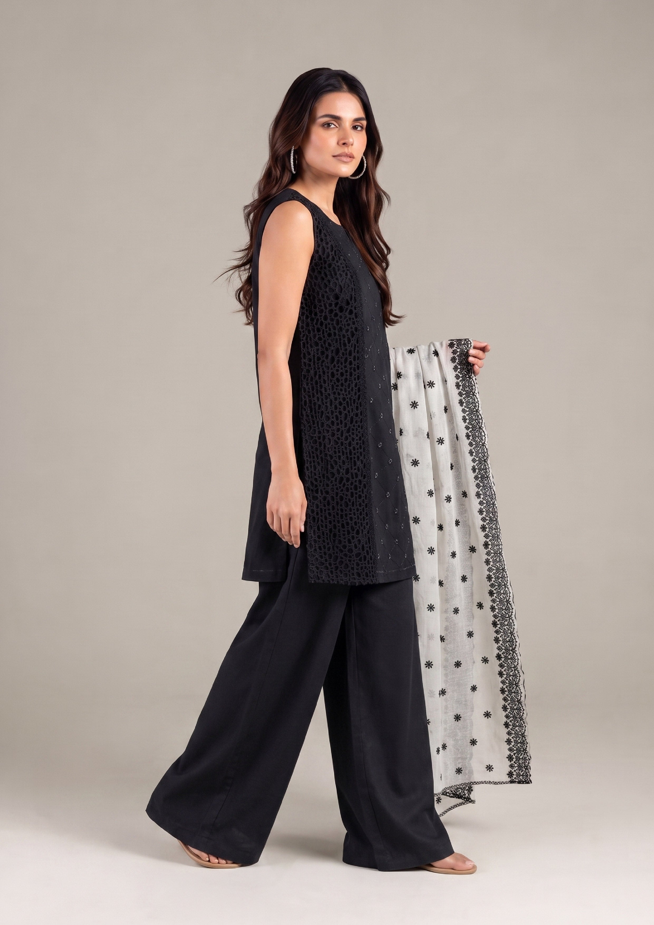 Model wearing a black sleeveless embroidered kurta with wide-leg palazzo pants and a white embroidered dupatta in a stylish side walking pose.