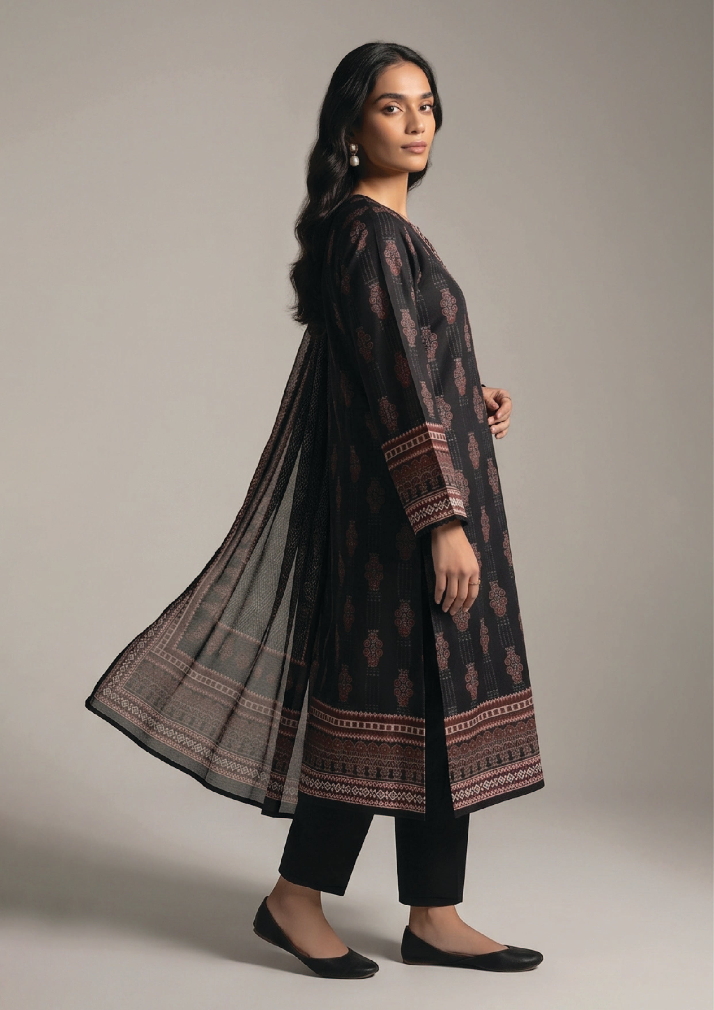 Seated model wearing Crimson Mosaic Noir black designer lawn suit with pearl earrings and patterned dupatta