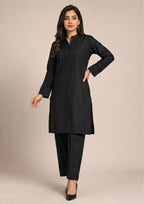 Model wearing Essential Pintuck Noir women co-ord set against wall showing long black shirt, button placket and tailored trouser