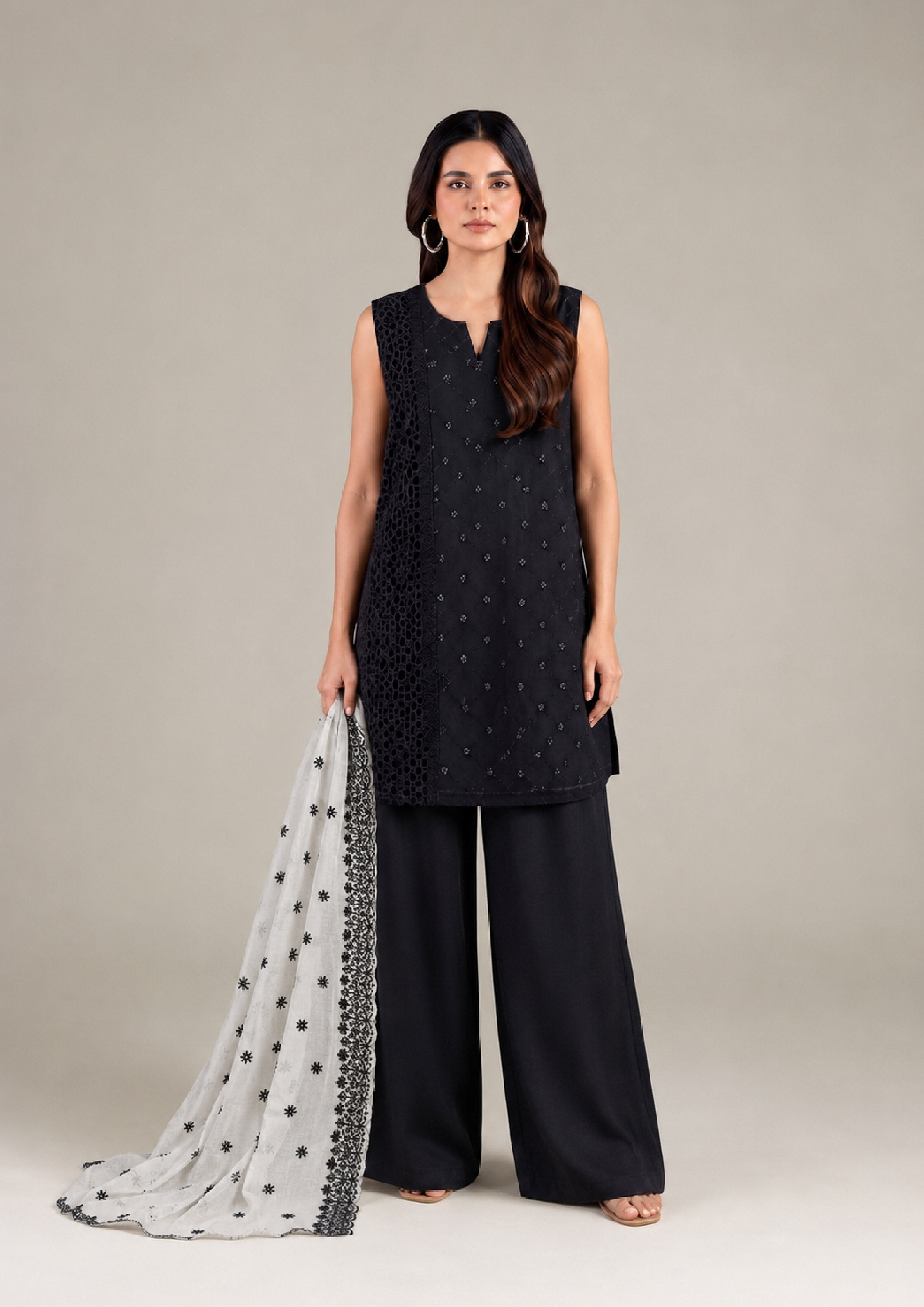 Full-length front view of a woman wearing a black embroidered sleeveless kurta, black palazzo trousers, and a white embroidered dupatta.