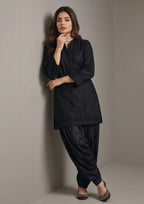 Seated view of Pleated Afghani Noir black coard suit for women with tonal pleated cuffs, matching Afghani shalwar and embroidered khussas