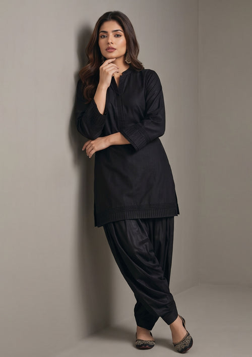 Seated view of Pleated Afghani Noir black coard suit for women with tonal pleated cuffs, matching Afghani shalwar and embroidered khussas
